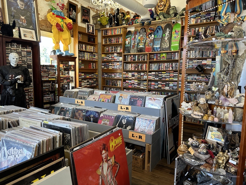 A record store with vinyl albums, skateboard decks, DVDs, and various collectibles displayed on shelves and tables.