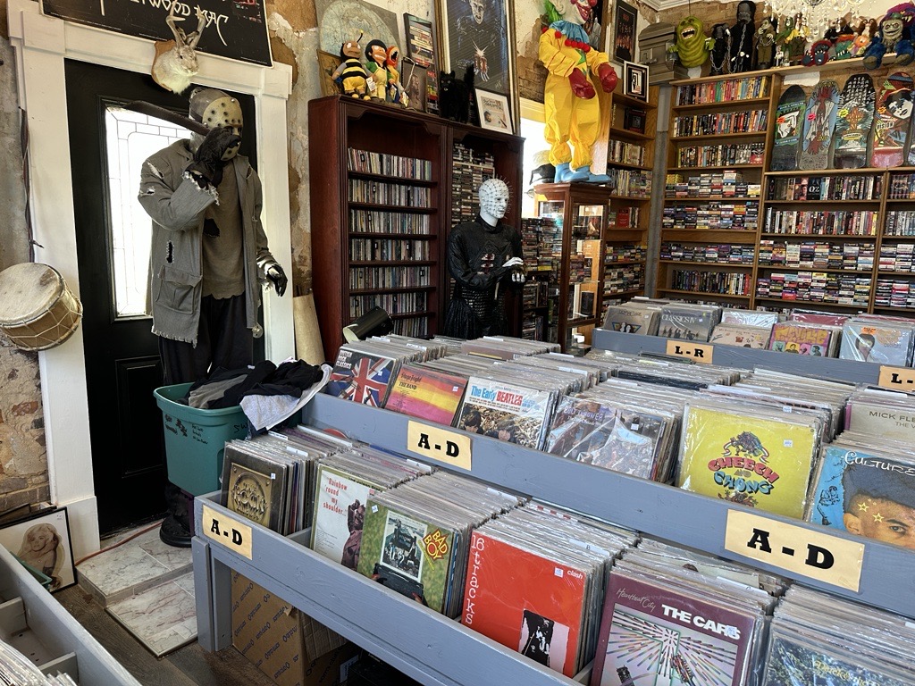 A vintage store with shelves of vinyl records categorized alphabetically. Mannequins in masks stand by the entrance. Walls are lined with CDs, and various decor items are visible.