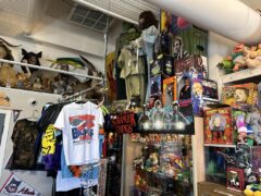 A cluttered store display features horror movie merchandise, including Chucky dolls, Stranger Things poster, collectible figures, T-shirts, and various vintage items on shelves and walls.