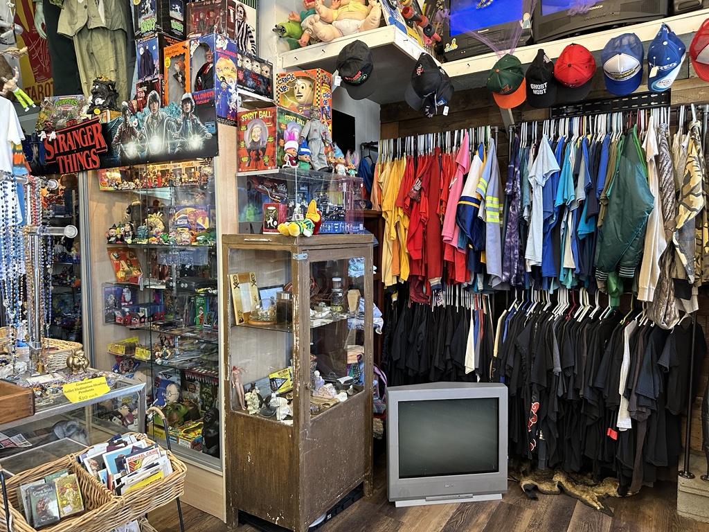 A vintage shop interior with a variety of merchandise including clothes on racks, hats on display, collectibles in glass cases, and an old television on the floor.