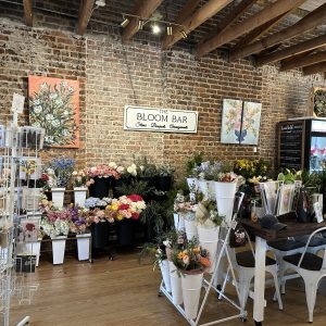 Flower shop interior with brick walls, floral arrangements in white buckets, and a sign reading "The Bloom Bar"—one of Acworth’s must-visit spots for the perfect Valentine’s Day. Wooden floors and ceiling beams create a cozy atmosphere.