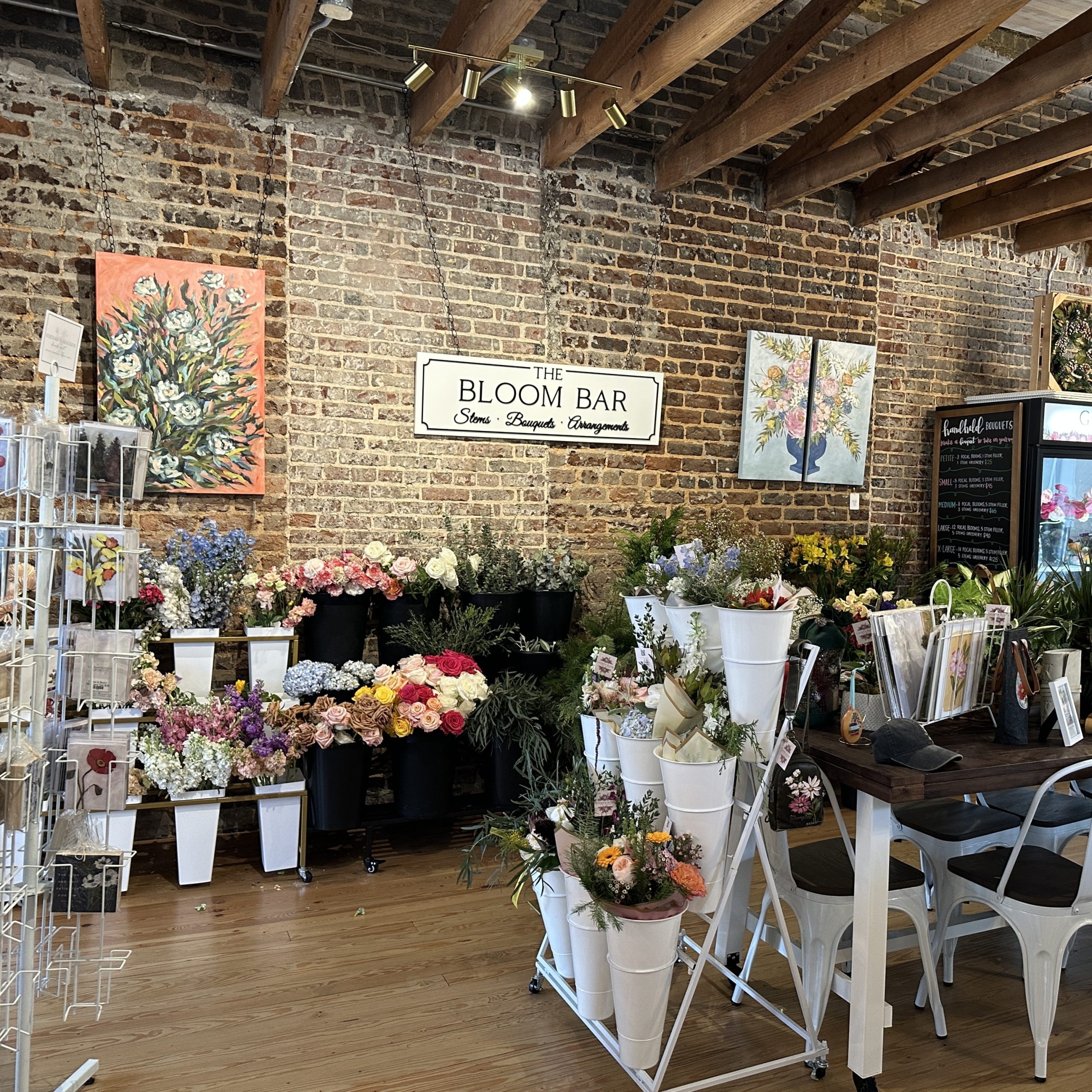 Flower shop interior with brick walls, floral arrangements in white buckets, and a sign reading "The Bloom Bar"—one of Acworth’s must-visit spots for the perfect Valentine’s Day. Wooden floors and ceiling beams create a cozy atmosphere.