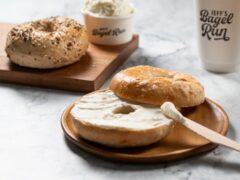 Bagels on a wooden plate, one sliced with cream cheese being spread. An everything bagel is on a cutting board. A cup and container with "Jeff's Bagel Run" branding are visible.