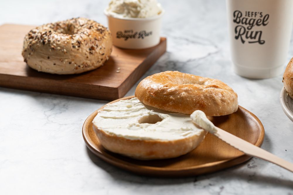 Bagels on a wooden plate, one sliced with cream cheese being spread. An everything bagel is on a cutting board. A cup and container with 