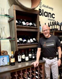 A man stands beside a wine rack filled with bottles, wearing a black "G Vino Market" shirt in a store.