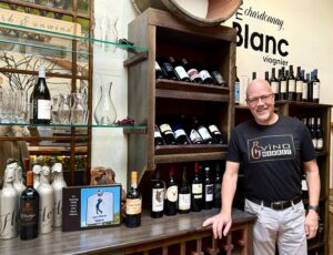 A man stands next to a wine display with various bottles and decanters. He wears glasses and a black T-shirt. Shelves hold red and white wine bottles in a well-lit setting.
