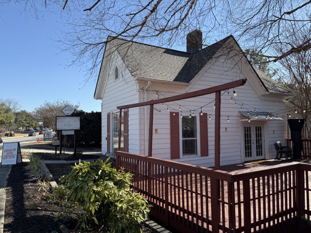 A small white house with red shutters and a wooden deck adorned with string lights. A sign nearby reads 