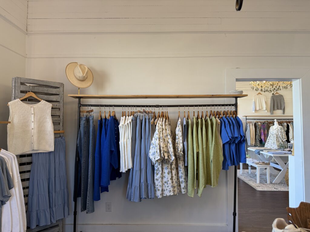 Clothing rack displaying various garments including shirts, jeans, and skirts in neutral and blue tones. A straw hat sits on the left end of the rack. Additional clothing visible in the background.