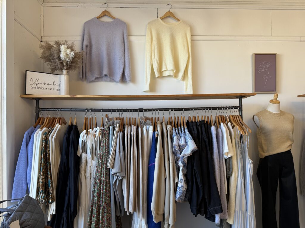 Clothing display in a store featuring sweaters, shirts, and dresses on hangers, with two sweaters hanging on the wall and a small framed sign on a shelf.