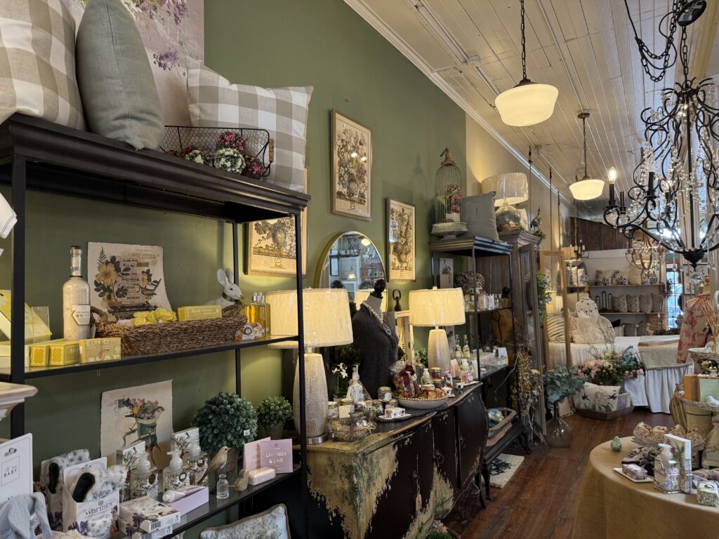 A cozy interior of a gift shop with green walls, showcasing various home decor items, lamps, cushions, and artwork. Shelves are filled with assorted gifts and decorative pieces.