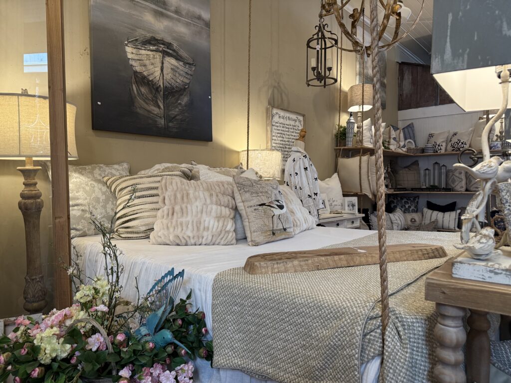 Cozy bedroom with a rustic theme featuring a bed with assorted pillows and a knit throw, surrounded by decorative lamps, flowers, and a painting of a boat on the wall.