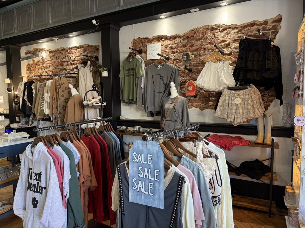 Clothing store interior with racks of assorted casual wear, including tops, hoodies, and shorts. Signs indicate a sale on several items. Exposed brick wall in the background.