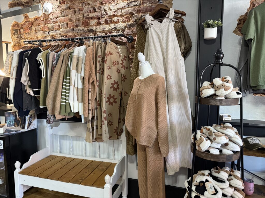 Clothing display showing various outfits on hangers, a beige mannequin, and a rack of slippers against a rustic brick wall.