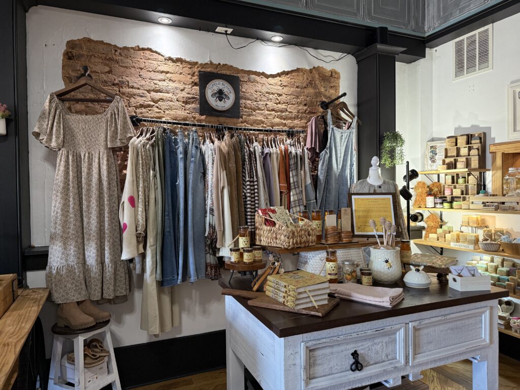 A boutique display featuring various dresses and shirts on a rack, along with a table showcasing honey jars, books, and soaps. Exposed brick wall and decorative items in the background.