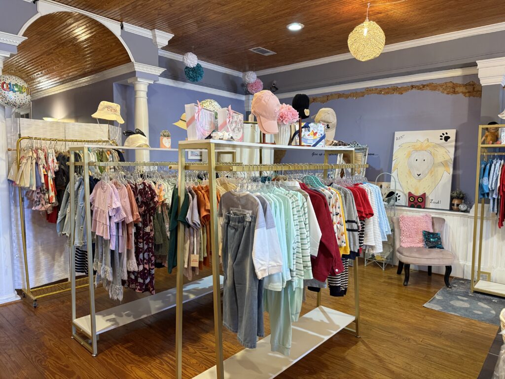 Children's boutique store interior with racks of colorful clothes, hats, and accessories. Wooden floors and decorative ceiling lights enhance the cozy atmosphere.