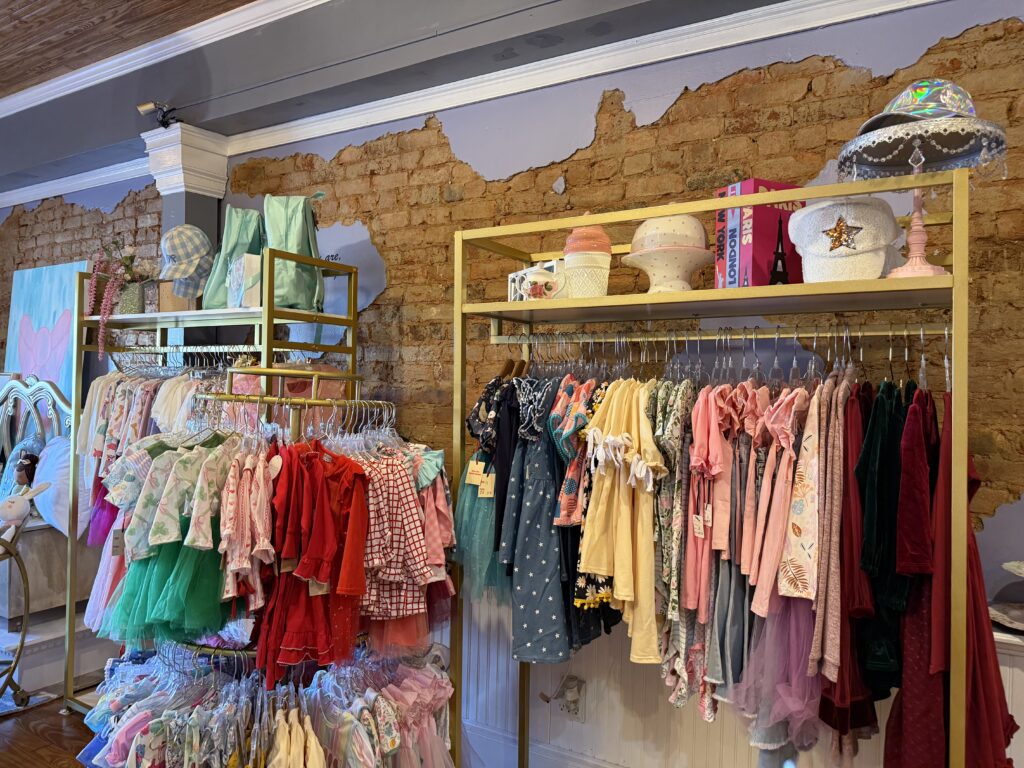 Children's clothing store with racks of colorful dresses, shirts, and hats against a rustic brick wall background.