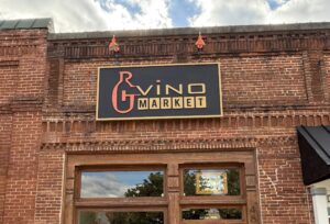 Brick storefront with a sign reading "RG Vino Market" above a wooden door and a bench outside.
