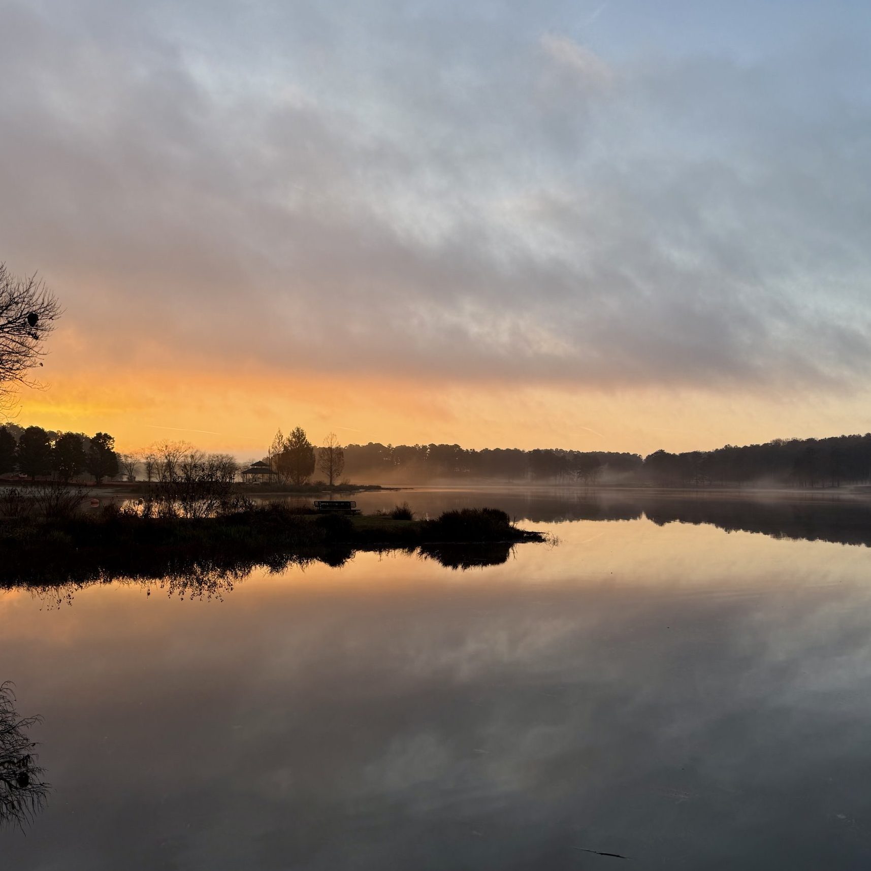 A calm lake at sunrise with orange light on the horizon, trees reflected in the water, and a wooden dock visible on the right—one of Acworth's must-visit spots for the perfect Valentine’s Day.