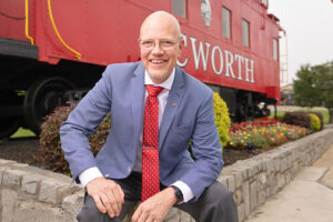 A man in a blue suit and red tie smiles in front of a red train car with "Worth" partially visible on it, surrounded by landscaping and flowers.