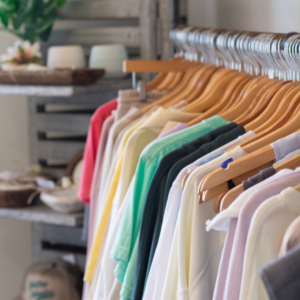 A row of neatly hung clothes in various colors on wooden hangers graces the well-lit retail store, adding charm akin to cozy winter combos that uplift your mood. Shelving with decor is visible in the background, enhancing the inviting atmosphere.