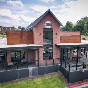 The historic brick building, featuring large windows and a gable roof marked with "1885," offers charming outdoor seating areas where guests can enjoy 6 Winter Combos for Fresh Food + a Good Mood in Acworth, surrounded by trees.