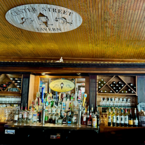 A well-stocked bar showcases various liquor bottles and glasses. A sign overhead with a decorative design reads "Center Street Tavern," setting a welcoming scene perfect for enjoying one of the 6 winter combos for fresh food + a good mood in Acworth. The ceiling features rustic wooden paneling.