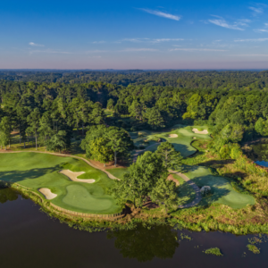 Aerial view of a golf course near Acworth, with manicured greens, sand bunkers, and lush trees beside a shimmering water body under a clear blue sky. It's one of five new reasons to put this charming location on your travel bucket list.