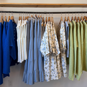 A clothing rack artistically showcases blue and green garments, perfect for winter, including cozy sweaters, chic blouses, and a stylish tiered skirt—all hanging on wooden hangers to uplift your mood like the fresh food in Acworth.