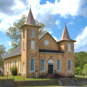 Historic brick church with two towers, arched windows, and a central entrance beckons you to Acworth. Surrounded by a metal fence, green trees, and grass under a partly cloudy sky—just one of the Five New Reasons to add Acworth to your travel bucket list.