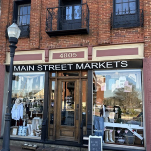 The brick storefront of "Main Street Markets," one of the 6 must-visit spots for the perfect Valentine's Day in Acworth, features large windows showcasing a variety of clothing and accessories. A lamppost stands to the left, and the building proudly displays the number 4805.