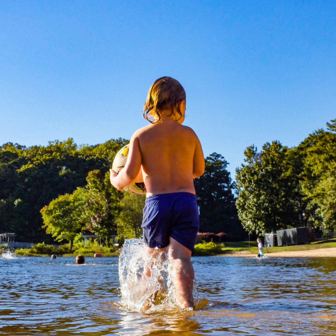 A child in swim shorts wades through the shallow water, clutching a ball against a backdrop of trees and clear blue sky—just one of the Five New Reasons to put Acworth on your Travel Bucket List.