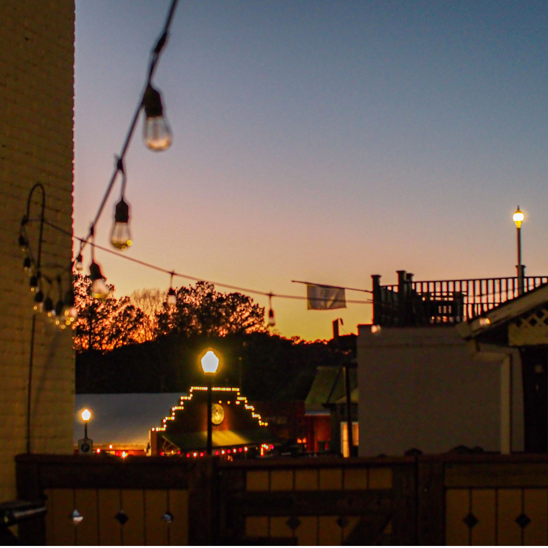 A string of lights hangs in the foreground with a sunset sky in the background, reminiscent of evenings you might find when exploring Acworth. Its charming rooftops and silhouetted lamp post at dusk offer just one of the five new reasons to put this enchanting destination on your travel bucket list.