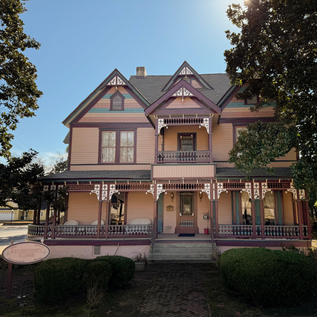 Victorian-style house with ornate details, pink and purple color scheme, surrounded by trees and shrubs.