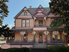 Victorian-style house with ornate details, pink and purple color scheme, surrounded by trees and shrubs.