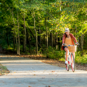 A woman rides a bicycle along a paved path through Acworth's wooded area, showcasing one of the five new reasons to put this charming spot on your travel bucket list. She sports a hat, an orange sweater, sneakers, and has a wicker basket attached to her bike's front.