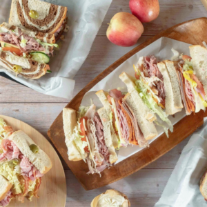 Assorted sandwiches with meats, lettuce, and cheese on a wooden platter, accompanied by apples and a small dessert on a wooden table—one of 6 Winter Combos for Fresh Food + a Good Mood in Acworth.