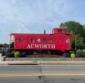 A red caboose with "ACWORTH" emblazoned on the side stands proudly as a landmark on a stone platform next to a street and lamp post, surrounded by lush greenery. Discover this charming spot and uncover five new reasons to put Acworth on your travel bucket list.