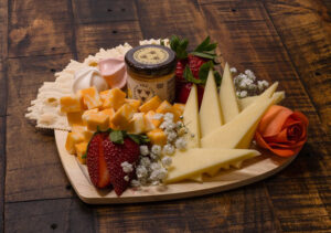 A wooden platter with various cheeses, crackers, strawberries, a small jar of dip, and a decorative rose-shaped garnish on a rustic wooden table.