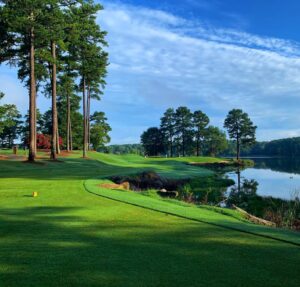 A serene golf course awaits in Acworth, offering neatly trimmed grass, tall pine trees, and a calm lake under a blue sky with scattered clouds—one of the five new reasons to put this charming town on your travel bucket list.