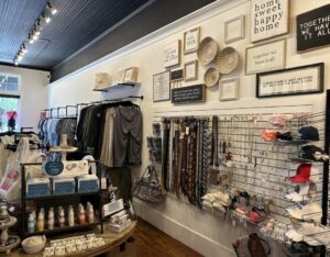 Discover a chic boutique that gives you five new reasons to put Acworth on your travel bucket list. Explore clothing racks, wall signs, belts, and accessories. A display table with skincare products sits against white walls and a wooden floor, adding to the inviting atmosphere.