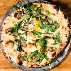 A wood-fired pizza topped with arugula, mushrooms, and a runny egg on a metal plate sits on a wooden table—one of the 6 winter combos for fresh food + a good mood in Acworth.