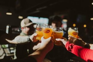 In a dimly lit setting, people clink glasses of beer with "Red Top" logos, celebrating the vibrant spirit that makes Acworth a must-visit. Discover five new reasons to put this charming town on your travel bucket list. Cheers to unforgettable adventures in Acworth!.