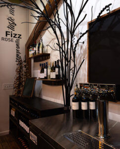 A stylish bar interior with wine bottles on shelves, a decorative metal tree, and a row of wine taps on a wooden counter.
