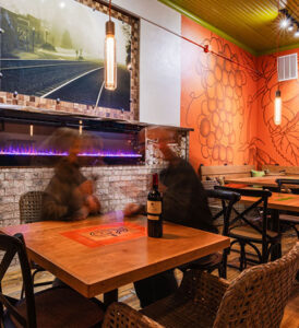 Shelves of assorted wine bottles above a cozy dining area with a mural and a wall-mounted fireplace.