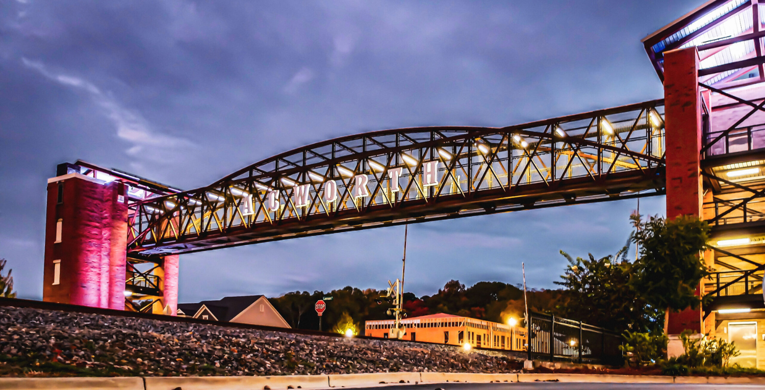 A lit pedestrian bridge with an arched design spans over a street at dusk, with trees and a building in the background, offering one of five new reasons to put Acworth on your travel bucket list.