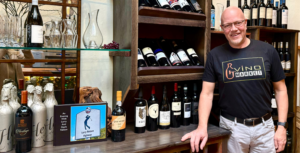 A man stands beside shelves of wine bottles and decanters in the great wine market in Downtown Acworth. A sign reads "An Evening With Larry Miller and Geoff Nelson," featuring an illustration of a golfer.