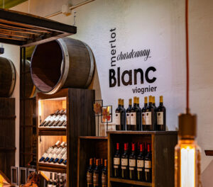 A wine display with wooden shelves showcases the best wine in Acworth, featuring a selection of bottled wines next to large barrels. Text on the wall lists types like Merlot, Chardonnay, Blanc, and Viognier. Warm lighting beautifully illuminates the area.