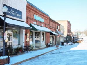 A snow-covered small town street in Acworth features brick storefronts, including a sandwich shop offering 6 Combos for Fresh Food + a Good Mood. Tables and chairs are set outside under green awnings, basking under the blue sky above.