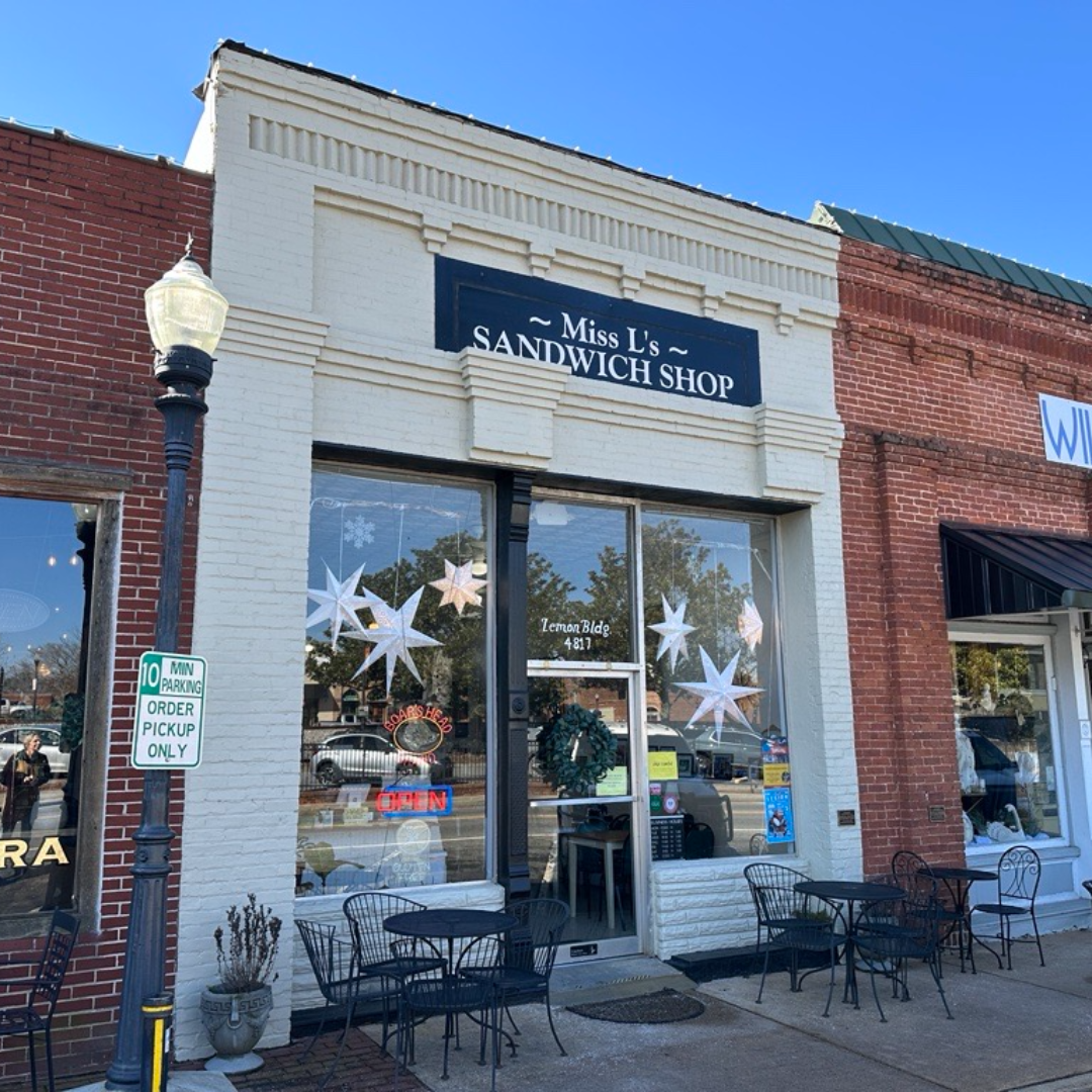 A sandwich shop with a sign reading "Miss L's Sandwich Shop" stands as one of the handful of reasons to put Acworth on your travel bucket list. The storefront features outdoor tables, chairs, and decorative stars in the window, offering a welcoming spot to enjoy a delicious meal.