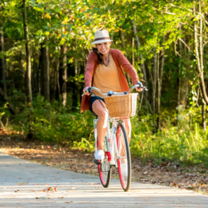 Smiling while riding a bicycle with a basket along Acworth's wooded path, wearing a hat and casual clothing—a perfect snapshot of why you should put this charming destination on your travel bucket list.